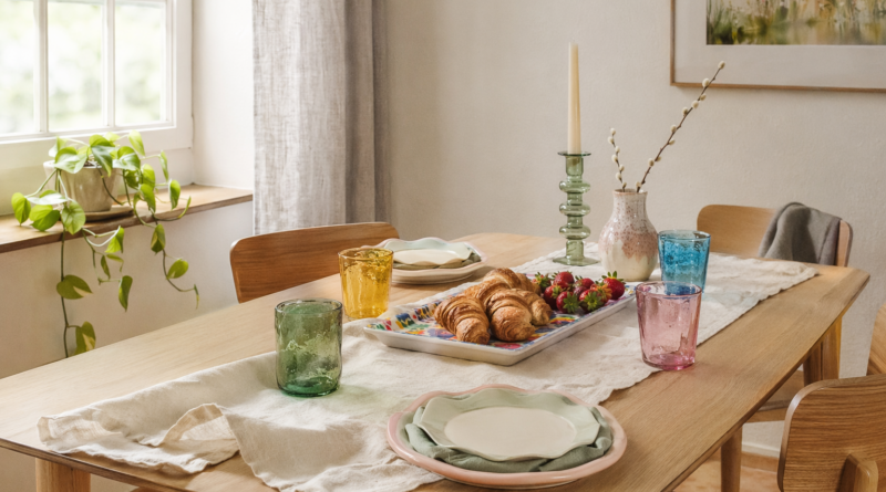 Весенняя сервировка коллекции TJÄRLEK: разноцветные стаканы, тарелки и подсвечник на светлом дубовом столе